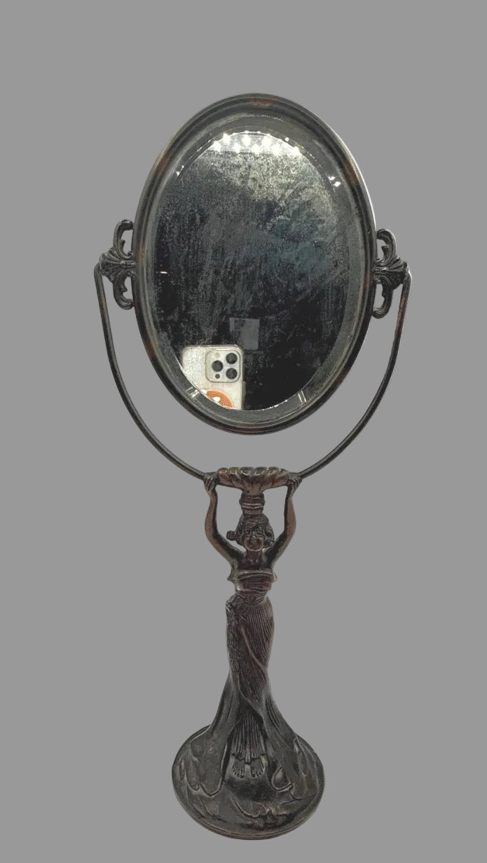 An antique-style oval vanity mirror with a dark metal frame and a sculpted female figure stand, shown against a plain white background.
