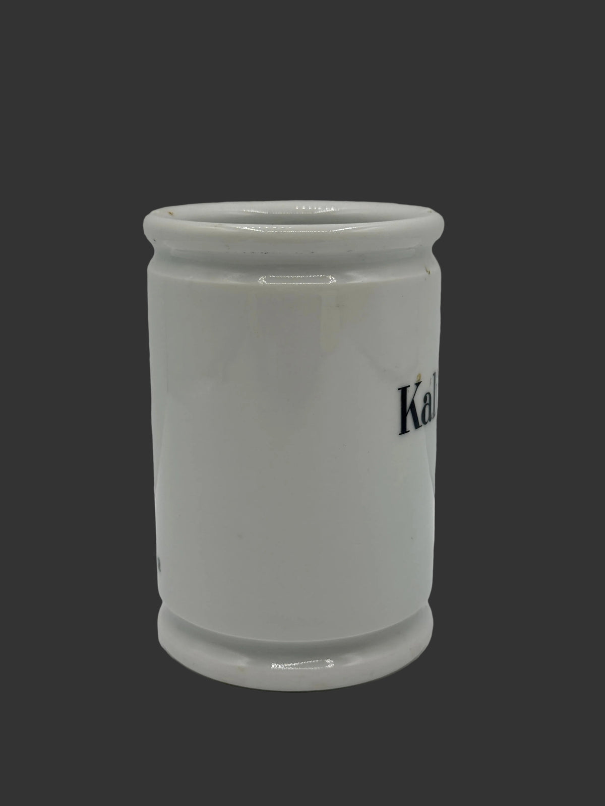 A white ceramic cup or small container with a cylindrical shape, featuring partial black text reading 'Ka' on the side, photographed against a plain light gray background.