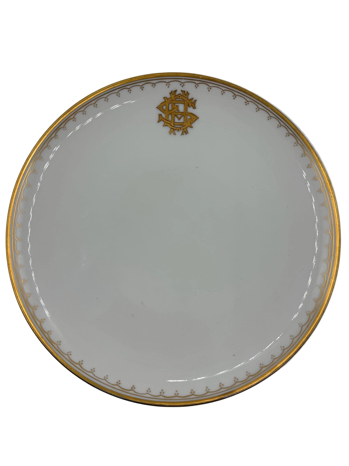 A round white porcelain plate with a gold rim, featuring a gold monogram crest at the top and a delicate gold pattern along the inner edge, shown against a neutral light gray background.