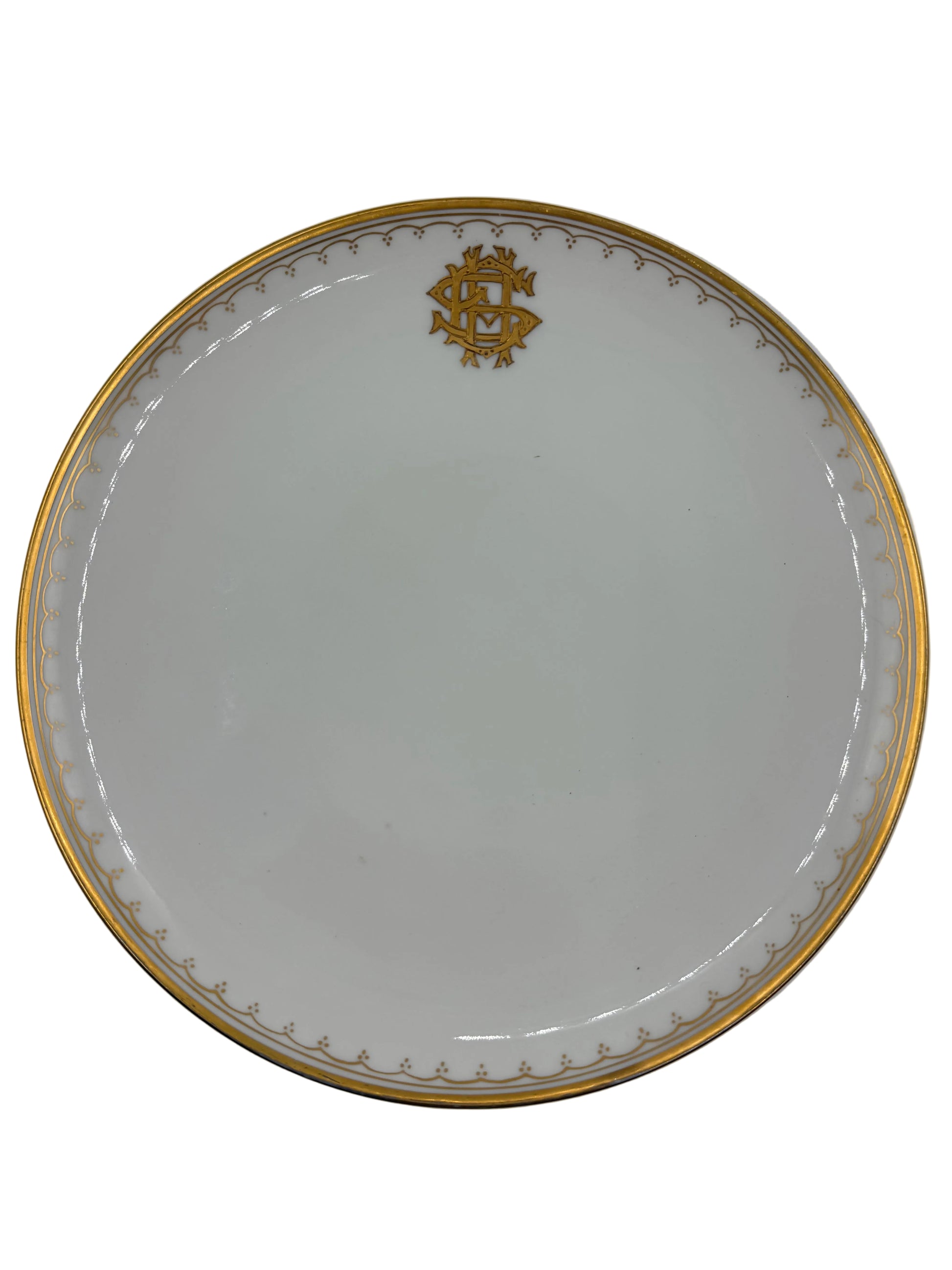 A round white porcelain plate with a gold rim, featuring a gold monogram crest at the top and a delicate gold pattern along the inner edge, shown against a neutral light gray background.