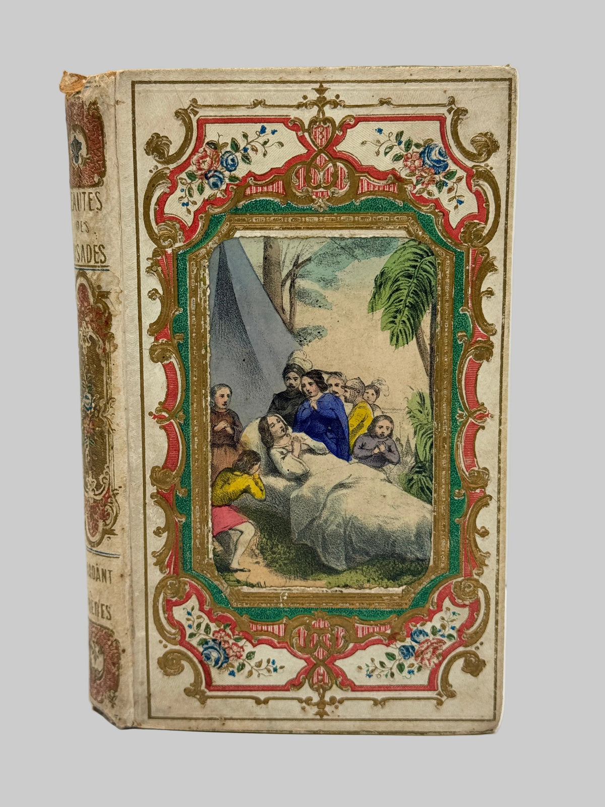 An antique hardcover book with a cream-colored cover, featuring an ornate red, green, and gold decorative border surrounding a central vintage-style illustration of a historical scene. The spine is visible on the left side.