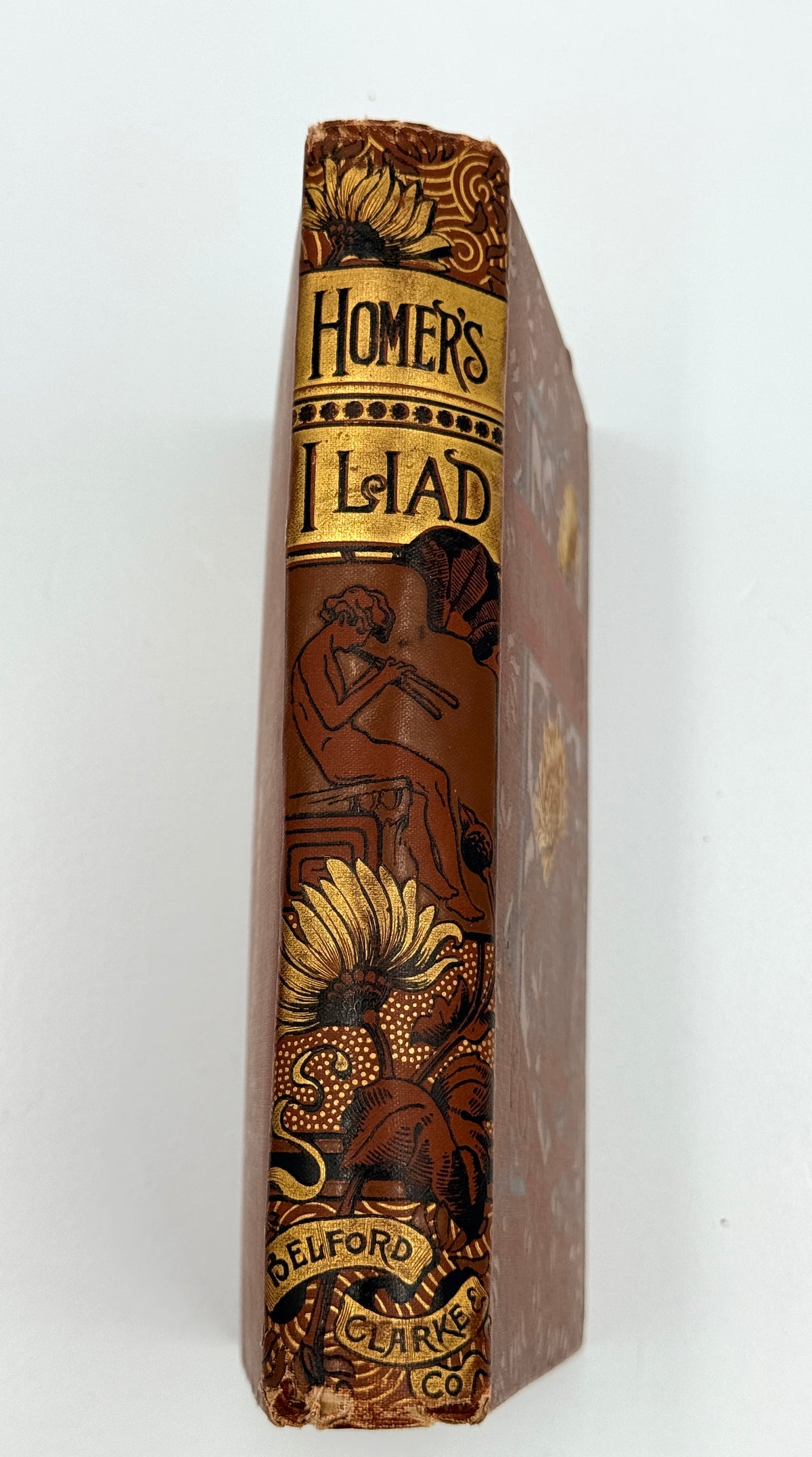 A vintage hardcover book, 'Homer's Iliad', standing upright against a plain white background. The book features an ornate brown and gold cloth spine with illustrations of a warrior and sunflowers, and the publisher's name 'Delford Clarke Co.