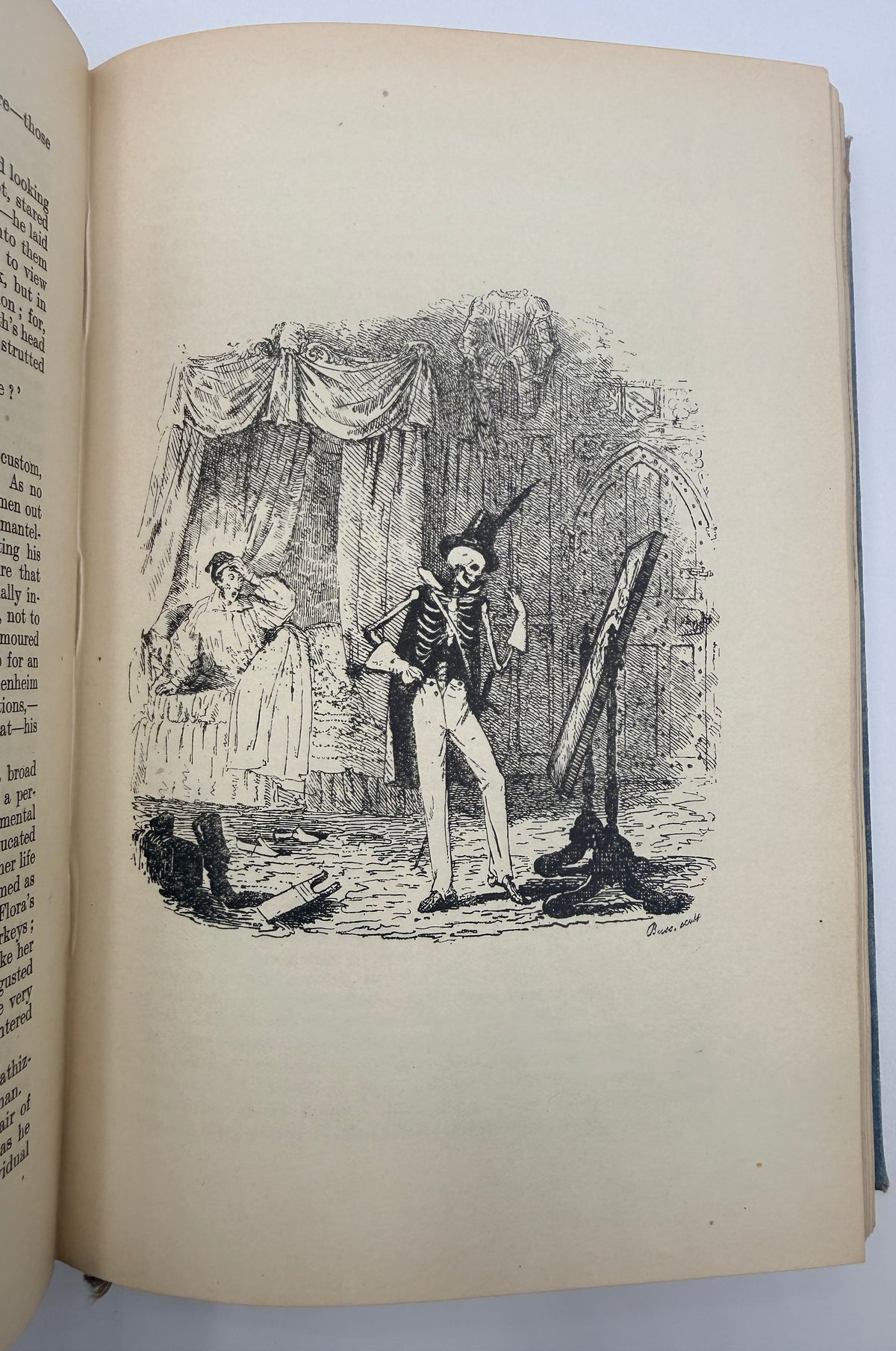 An open vintage book showing a black ink illustration on aged, cream-colored paper. The illustration depicts a historical scene with a skeleton-like figure standing before a mirror, accompanied by a seated figure and a dog, while the left page contains printed text.