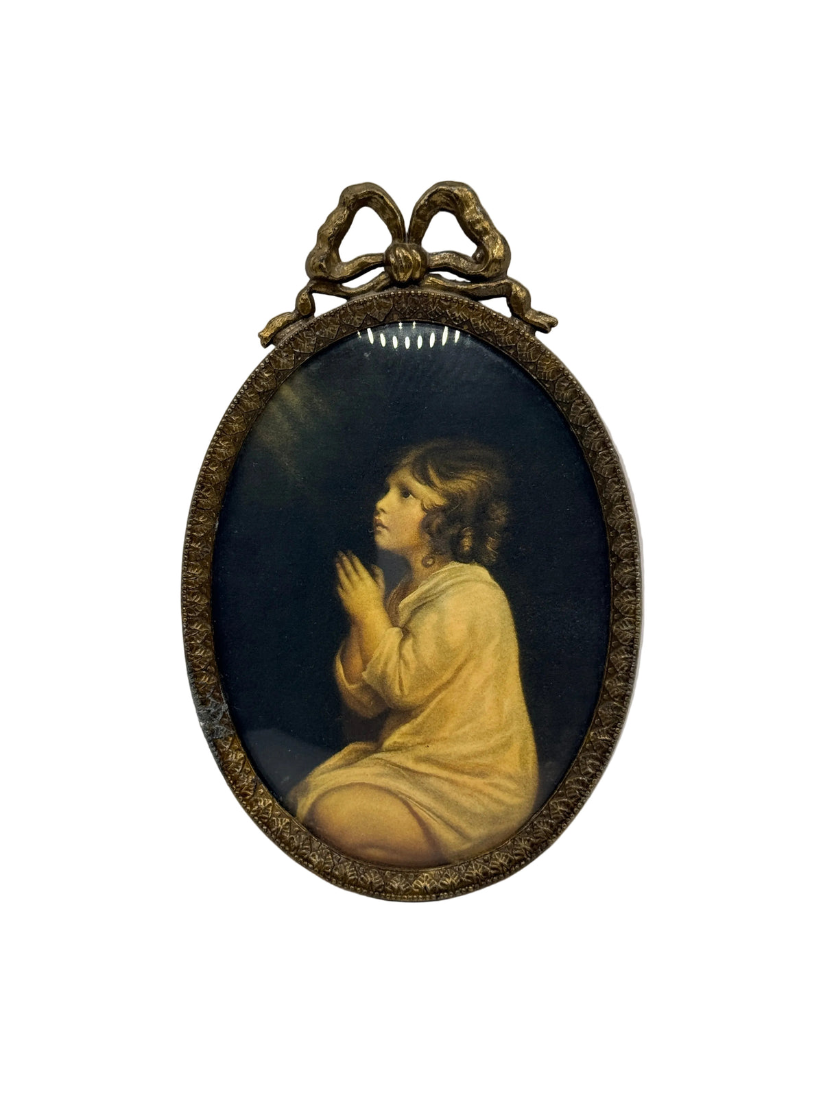 An oval religious wall art piece featuring a classical painting of a kneeling, praying child in a golden robe against a dark background, surrounded by an ornate bronze frame with a decorative bow at the top.