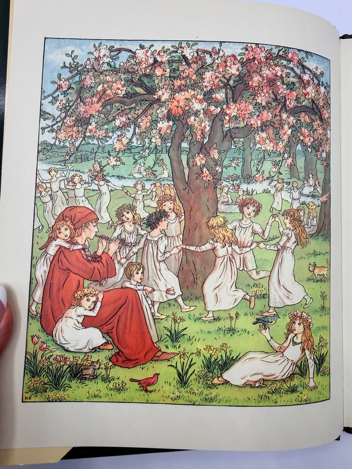 An open vintage book or art print showing a whimsical illustration of children playing under a large blooming tree in a grassy park. A hand is visible holding the left page edge.