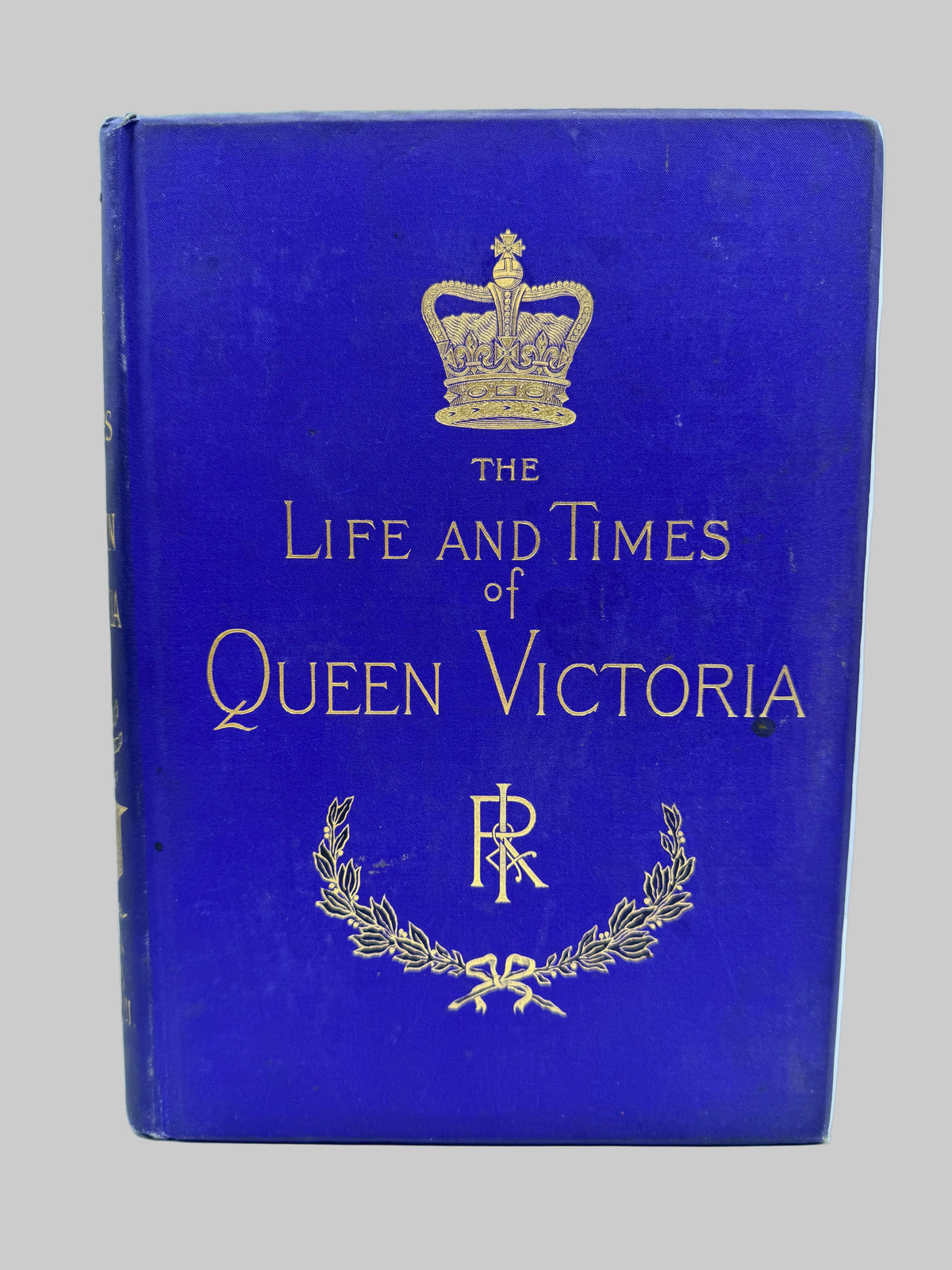 A vintage hardcover book titled 'The Life and Times of Queen Victoria' with a deep blue cover and gold foil detailing, including a crown, royal letters, and laurel wreath, set against a light blue background.