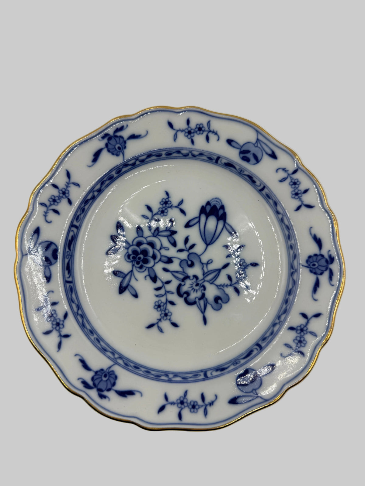 A scalloped ceramic plate with a blue floral pattern and gold trim, photographed from directly above on a plain white background.