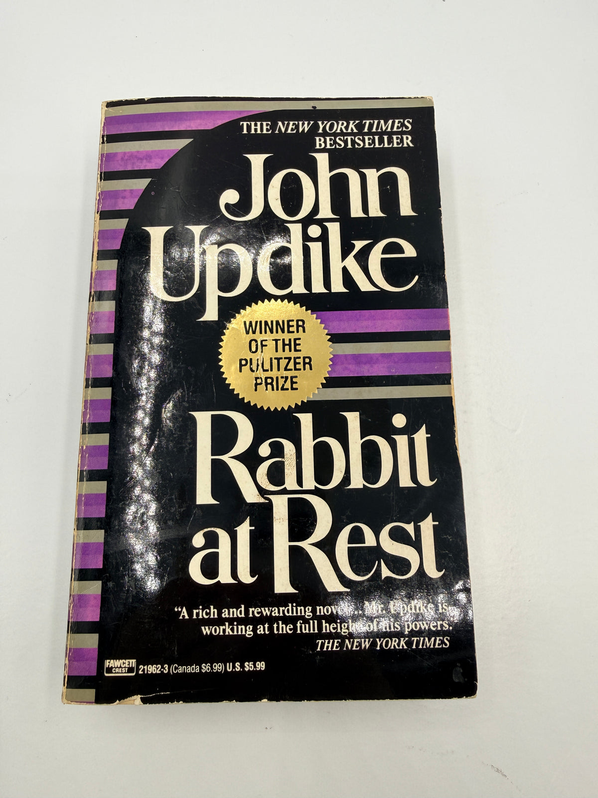 A vintage paperback book of John Updike's 'Rabbit at Rest' on a plain off-white background. The cover is black with purple stripes, cream-colored title text, and a gold Pulitzer Prize badge