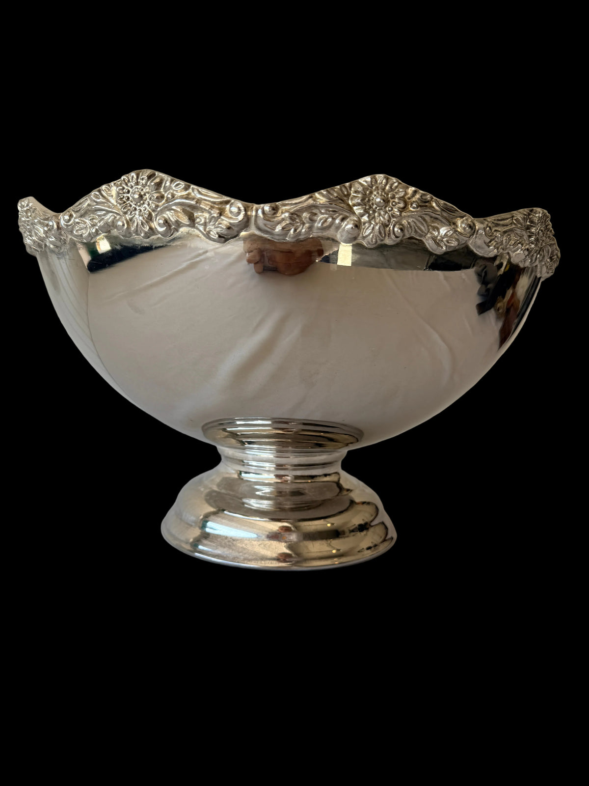 A polished silver pedestal bowl with an ornate, decorative floral and scrollwork rim, displayed on a white fabric surface against a white paneled wall.