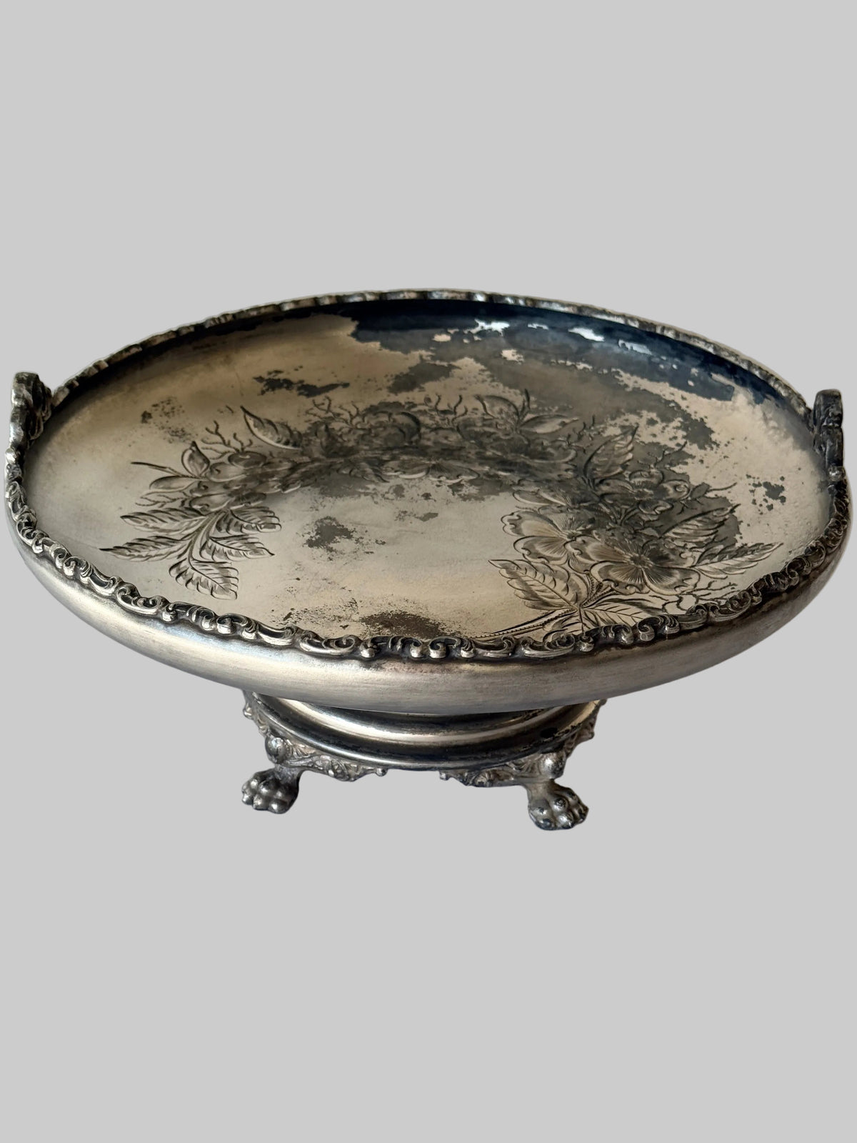 An antique silver pedestal bowl with floral engravings on the interior, featuring decorative side handles and a three-legged stand, set against a solid gray background.