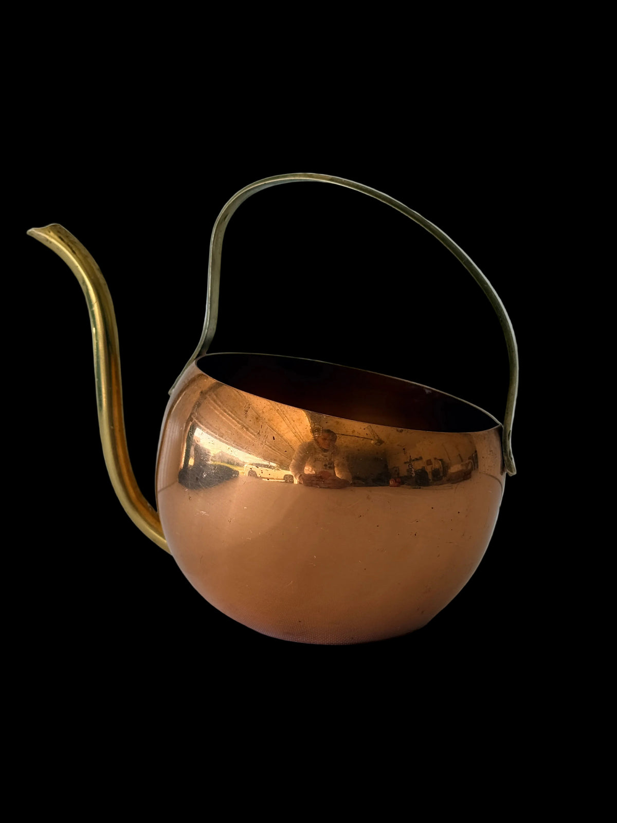 A polished copper teapot with a brass spout and handle, resting on a white surface against a white paneled background.