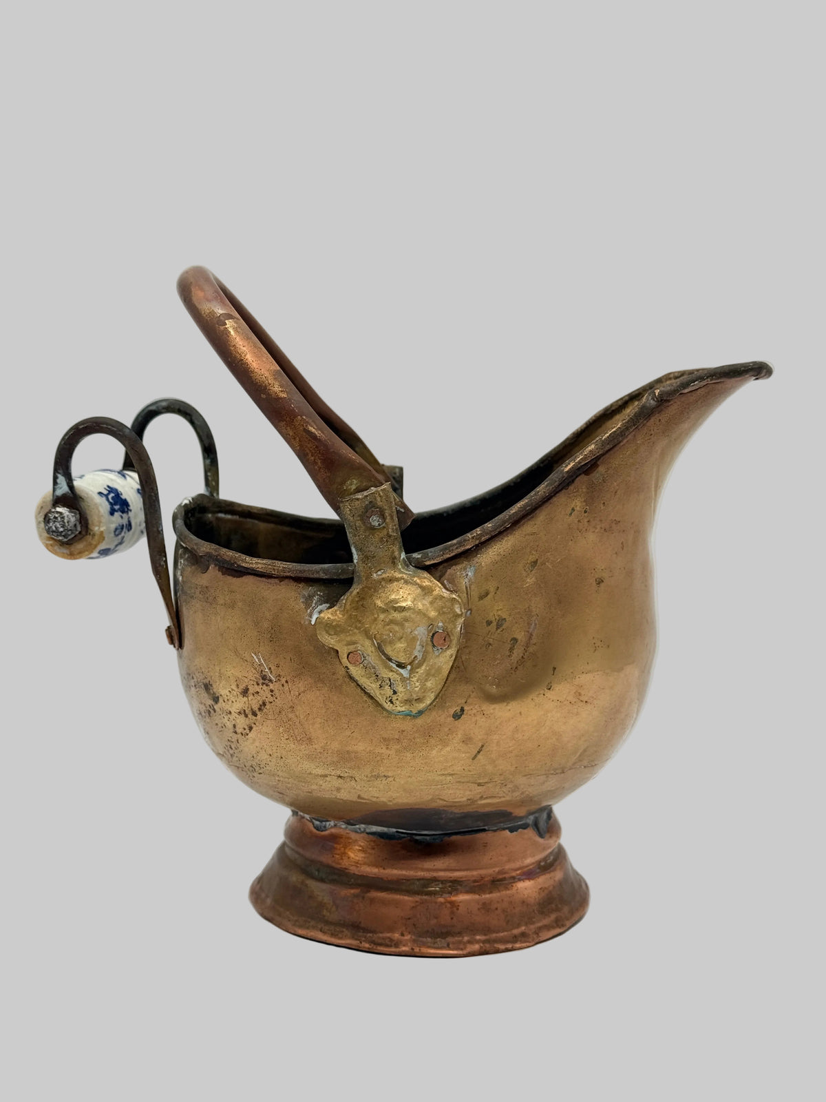 An antique brass or bronze wall-mounted pitcher with a curved spout and a hanging small white ceramic tag, shown against a plain light gray background.
