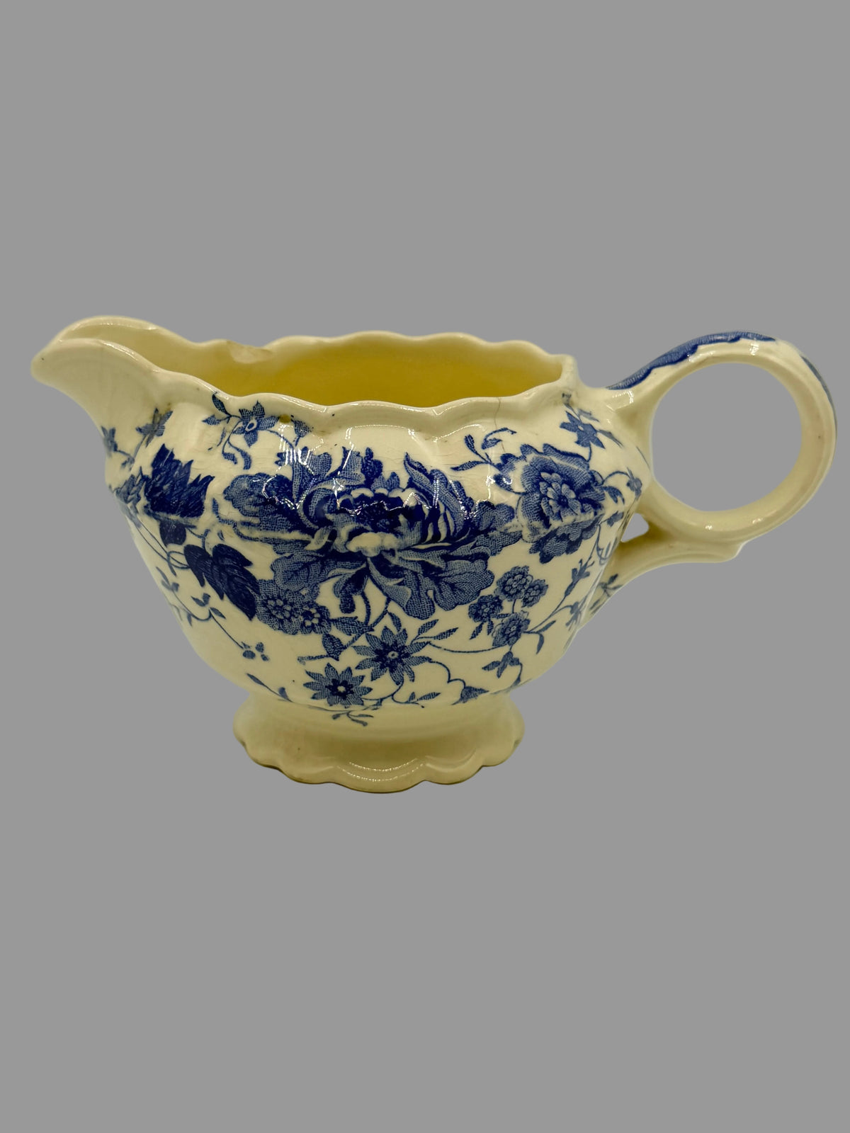 A vintage cream-colored ceramic creamer or sauce pitcher with a blue floral transfer pattern, featuring a fluted rim, scalloped handle, and footed base.