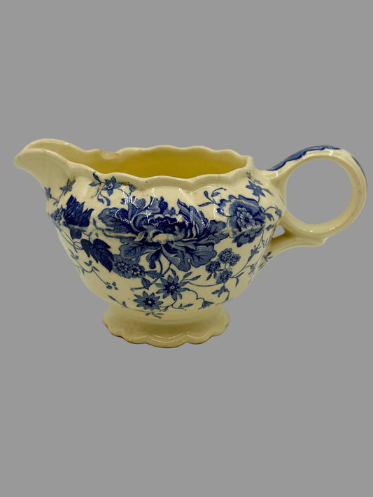 A vintage cream-colored ceramic creamer or sauce pitcher with a blue floral transfer pattern, featuring a fluted rim, scalloped handle, and footed base.
