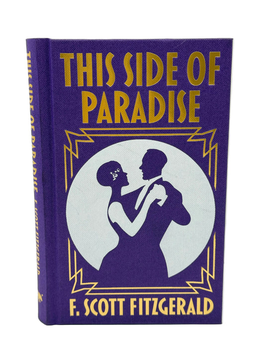 A hardcover book of F. Scott Fitzgerald's 'This Side of Paradise' with a purple cloth binding and gold lettering, featuring a white circular illustration of a dancing couple on the front cover.