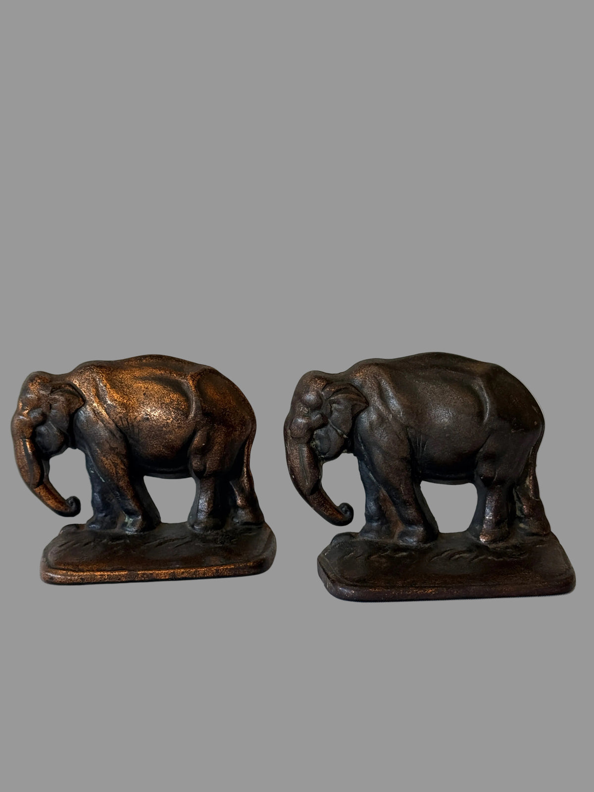 A pair of small elephant figurines with a dark bronze finish, standing on rectangular bases against a white paneled wall.