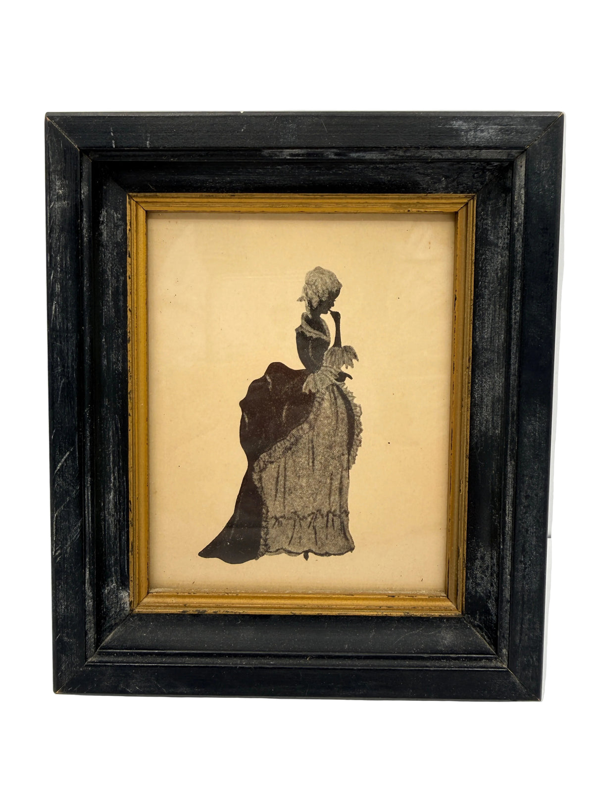 A framed vintage silhouette artwork of a woman in a long dress, mounted on a beige background with a gold inner frame and a distressed black outer frame, shown against a white wall.