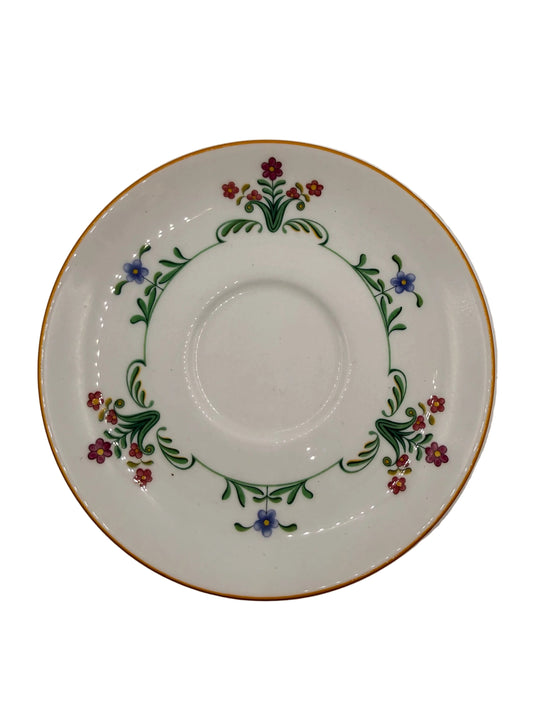 A white porcelain saucer decorated with a delicate floral border featuring green leaves, red and purple flowers, and a thin gold rim, shown on a plain light gray background.