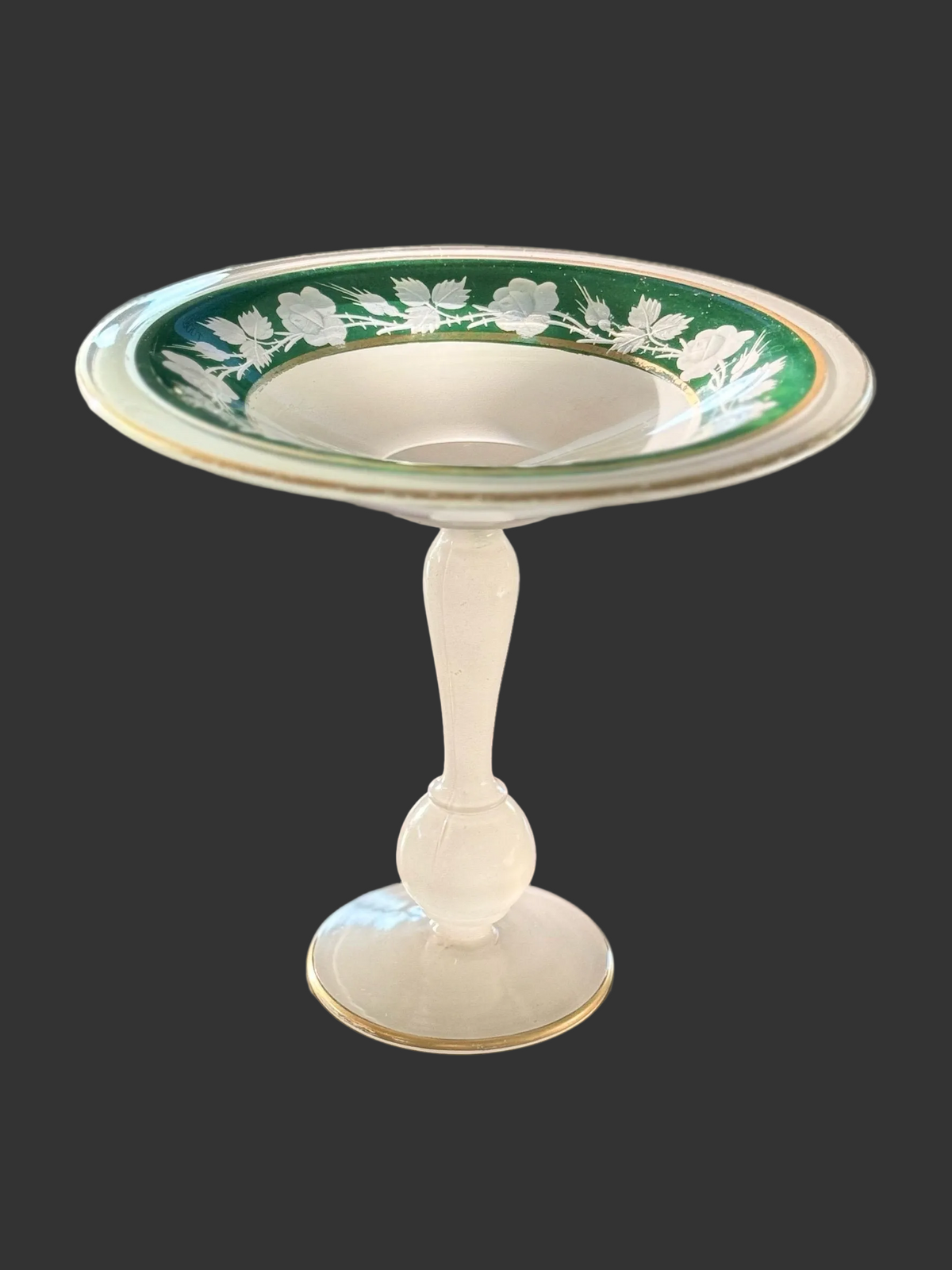 A vintage white glass pedestal bowl with a green and gold floral pattern around the rim, displayed on a dark wooden table.