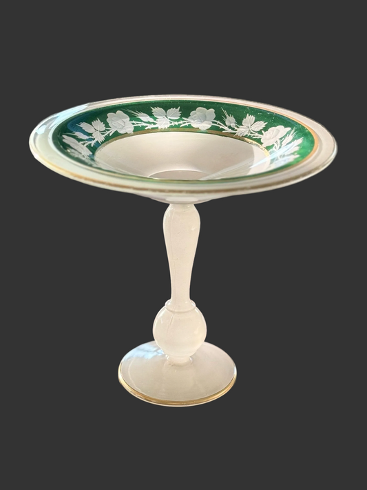 A vintage white glass pedestal bowl with a green and gold floral pattern around the rim, displayed on a dark wooden table.