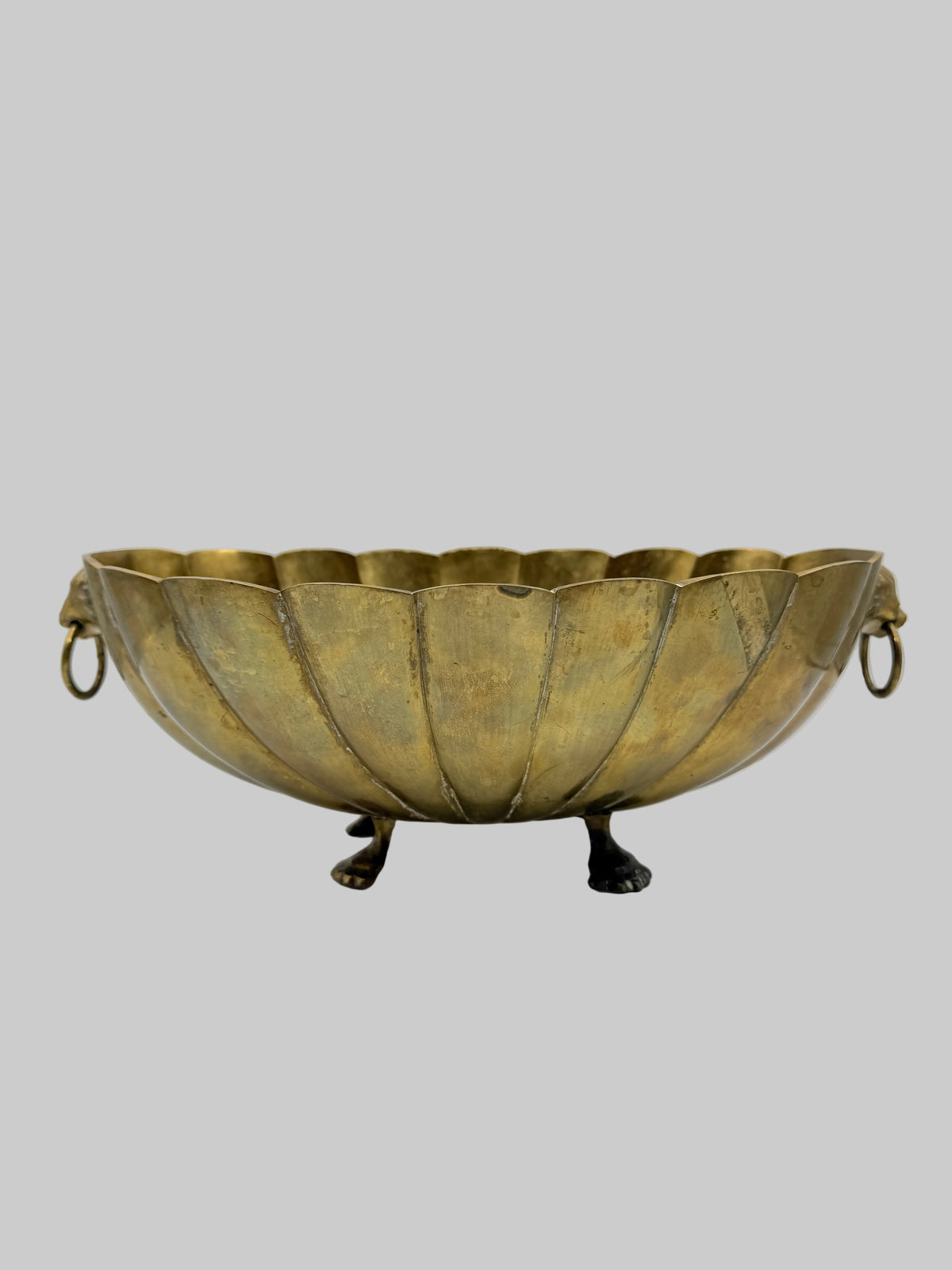 An antique-style brass pedestal bowl with a scalloped rim, lion head ring handles, and three small lion feet legs, shown on a white studio background.