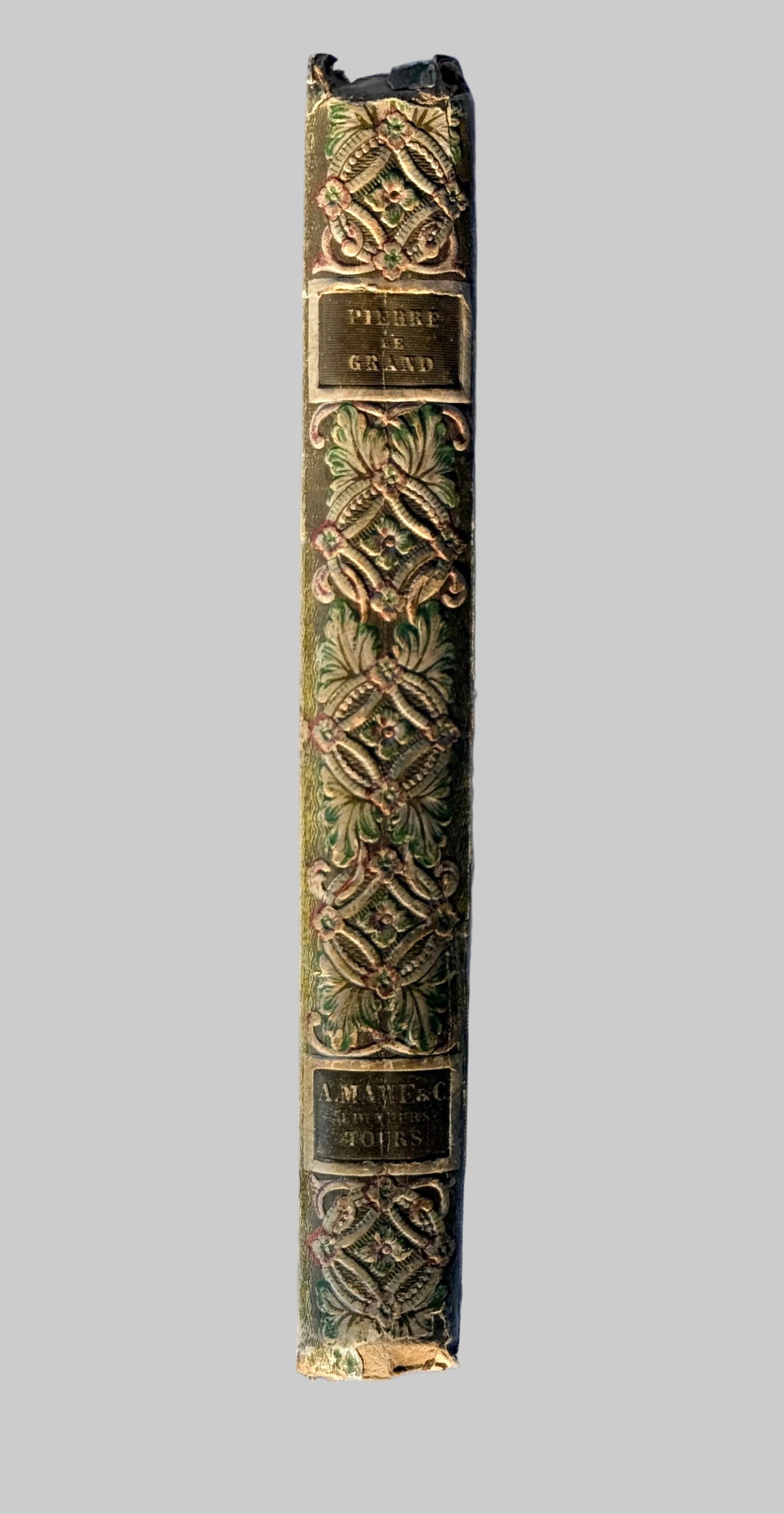 A close-up of an antique book's spine, featuring ornate gold embossed floral and geometric patterns on a dark green cloth, laid flat on a wrinkled light blue fabric background.