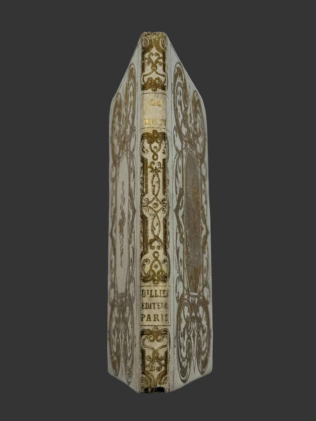 An antique octagonal book standing upright, showing its ornate gold-foiled spine and decorated cream-colored cover against a plain white background.