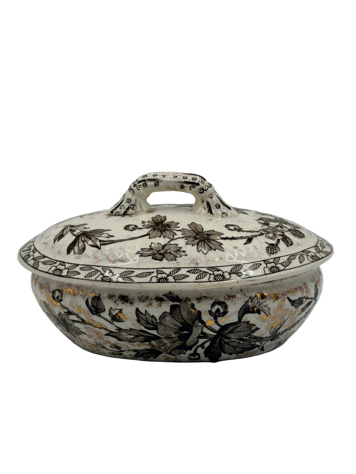 An antique ceramic lidded tureen with floral patterns in black, brown, and gold on an off-white background, photographed against a plain light gray wall.