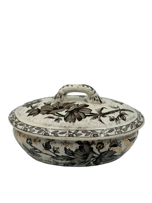 A vintage ceramic tureen with its lid on, featuring a dark brown floral and leaf pattern on an off-white glazed base, photographed against a plain light gray background.