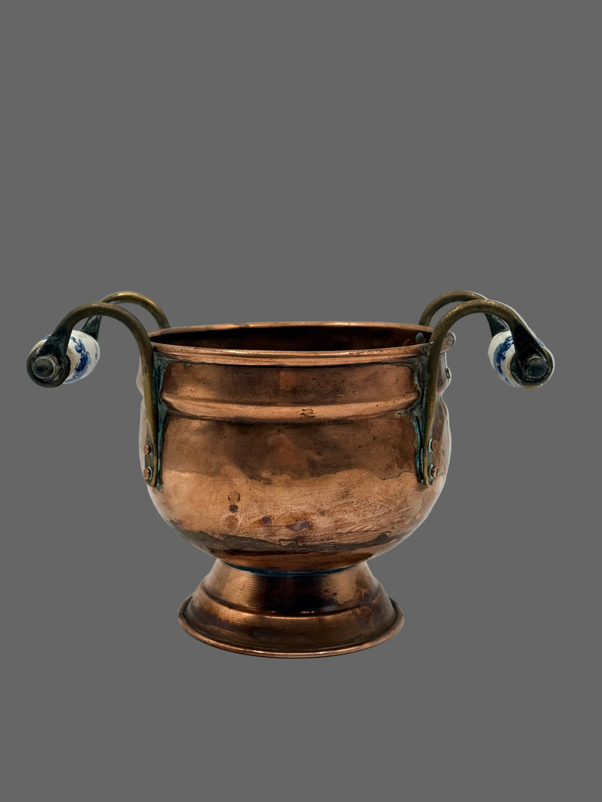 A vintage copper footed pot with curved handles and blue-and-white ceramic end fittings, photographed on a light gray background.