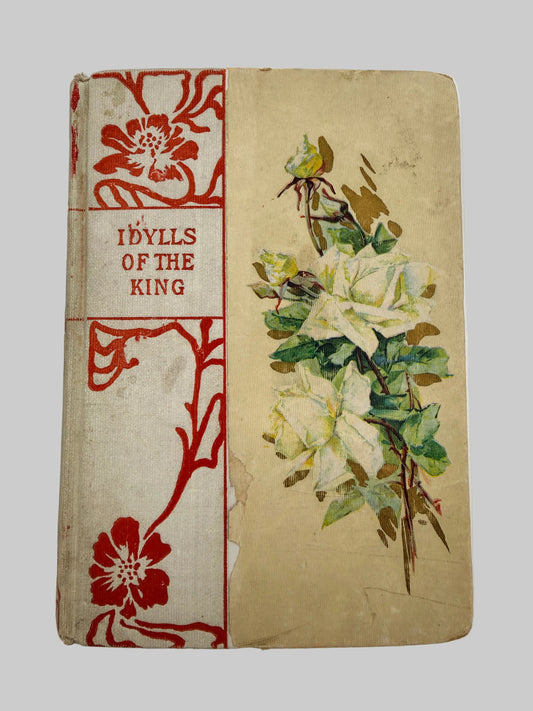 Vintage hardcover book titled 'Idyls of the King' with a beige cover, red floral border design on the spine, and an illustration of white flowers with green leaves on the front.