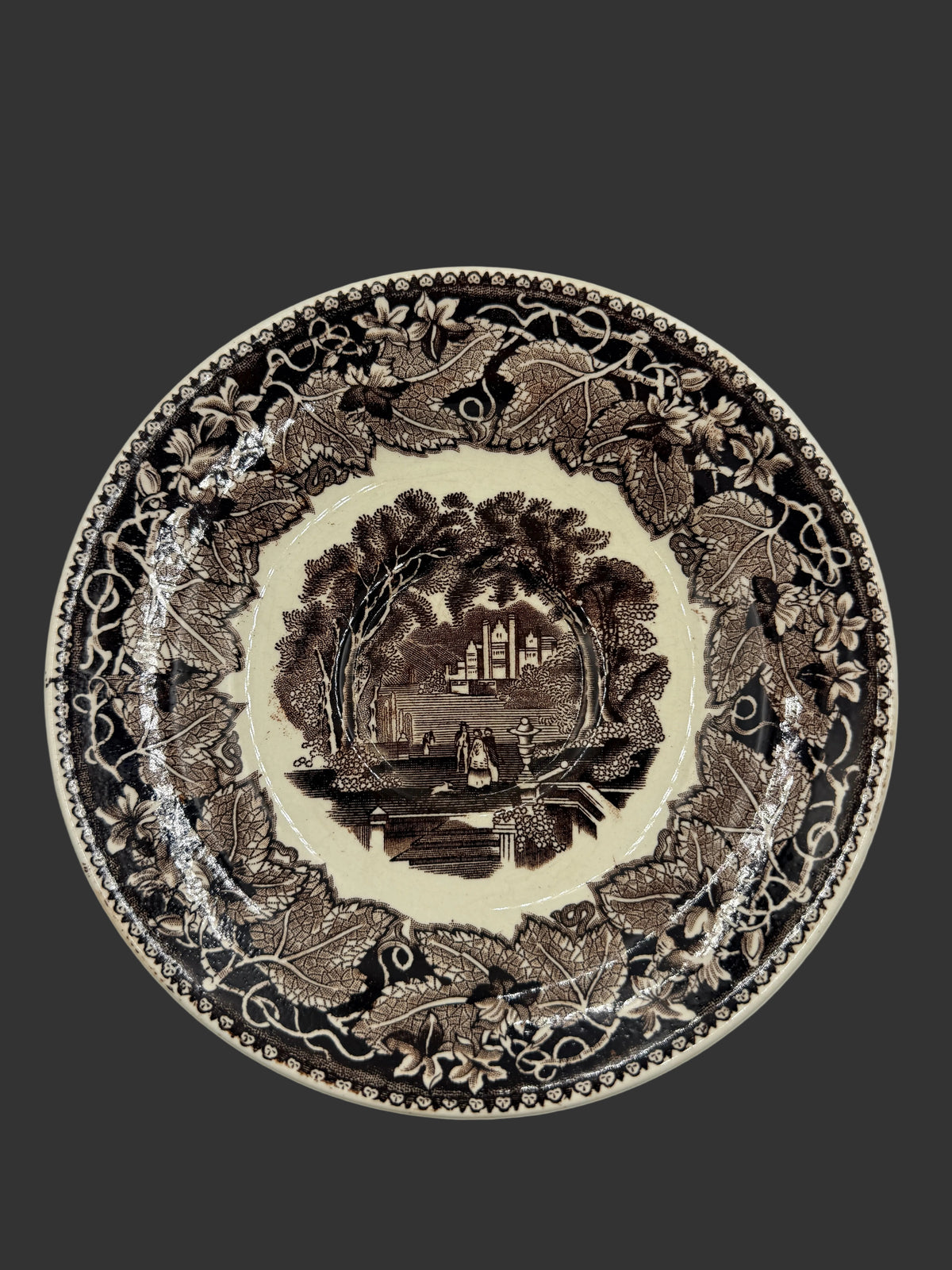 A round ceramic plate with a dark brown sepia transfer print featuring a central landscape scene and an ornate border of leaves and birds on a light cream background.
