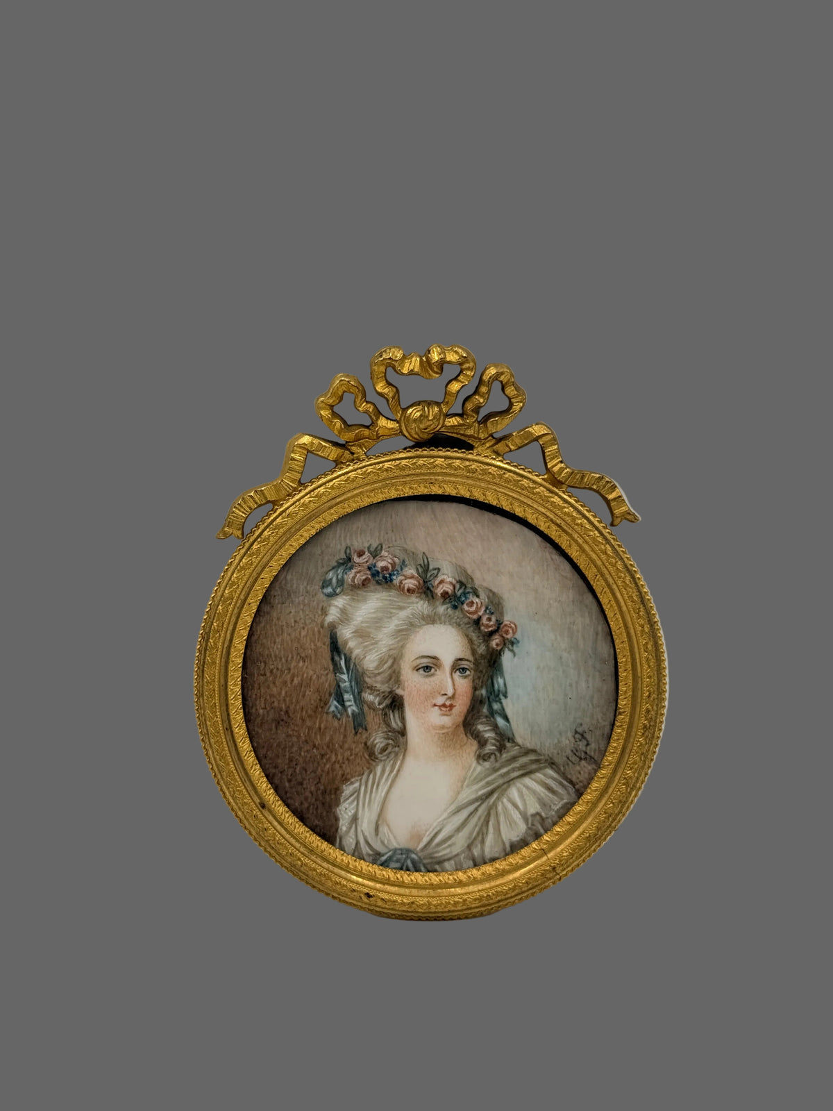 An antique-style oval gold-framed miniature portrait painting of a woman with a floral hairpiece, displayed against a plain light gray background.