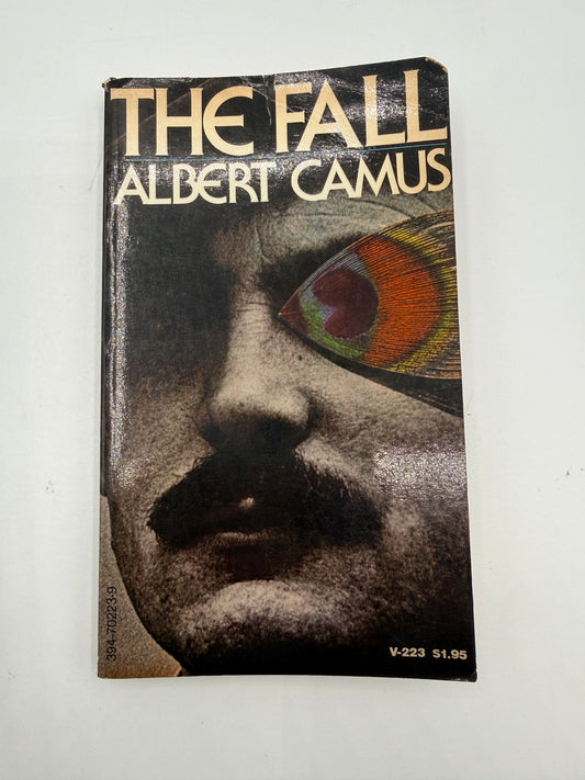 The Fall by Albert Camus paperback book cover, featuring a close-up portrait of a face with one eye replaced by a colorful peacock feather, photographed on a light off-white background.