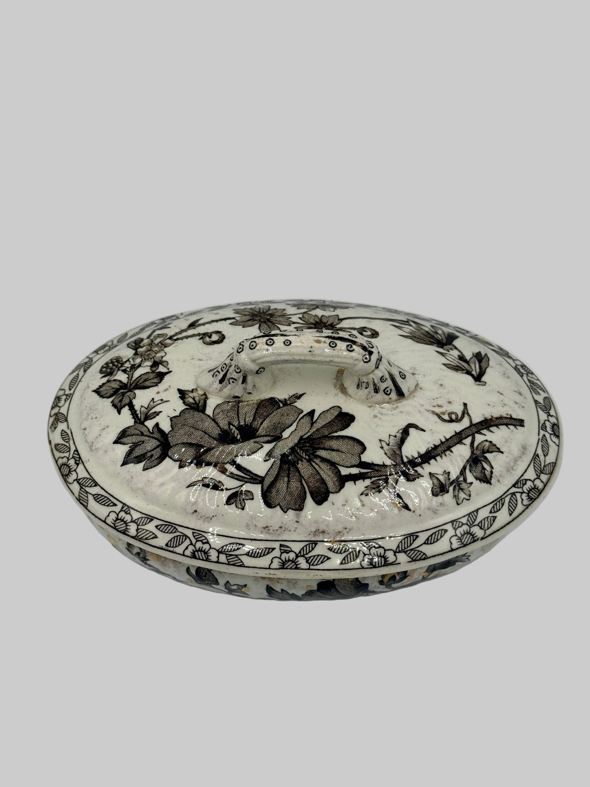 An oval ceramic wall sconce with a floral and leaf pattern, featuring a textured white background and dark gray detailing, shown against a plain white wall.